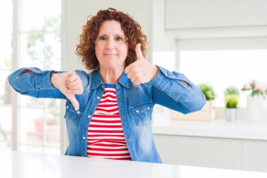 Evde kot ceket giyen kıvırcık saçlı orta yaş yaşlı kadın başparmak yukarı ve aşağı yapıyor, anlaşmazlık ve anlaşma ifade. Çılgın çatışma