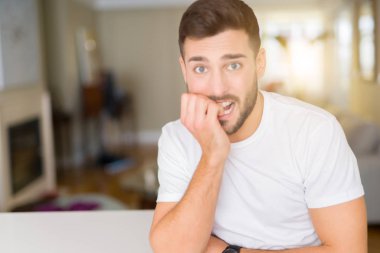 Evde rahat beyaz t-shirt giyen genç yakışıklı adam vurguladı ve ağız ısırma çivi elleri ile gergin bakıyor. Anksiyete sorunu.