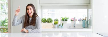 Evde beyaz masada oturan güzel genç kadının geniş açılı resmi başarılı bir fikirle parmağını yukarı doğru gösteriyor. Çıktı ve mutlu. Bir numara.