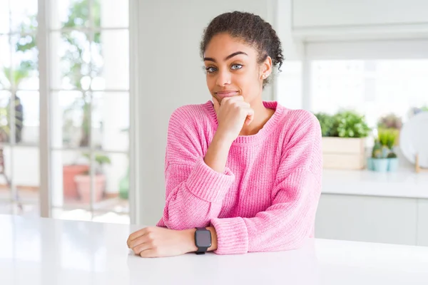 Beautiful african american woman with afro hair wearing casual pink sweater looking confident at the camera smiling with crossed arms and hand raised on chin. Thinking positive.