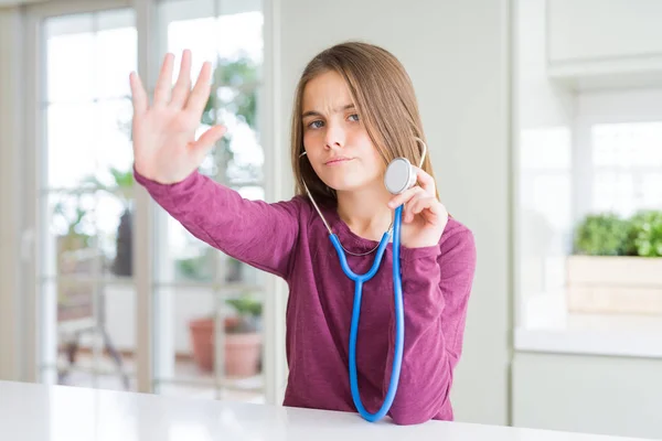 Beautiful young girl kid checking health using medical stethoscope with open hand doing stop sign with serious and confident expression, defense gesture