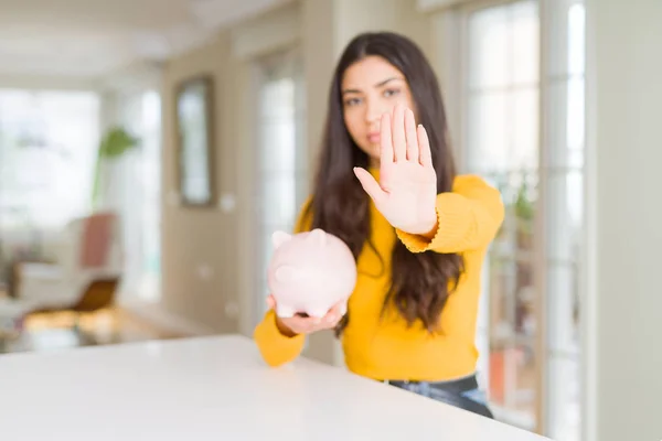 Young woman holding piggy bank with open hand doing stop sign with serious and confident expression, defense gesture