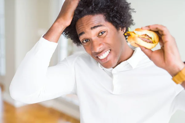 African American hungry man eating hamburger for lunch stressed with hand on head, shocked with shame and surprise face, angry and frustrated. Fear and upset for mistake.