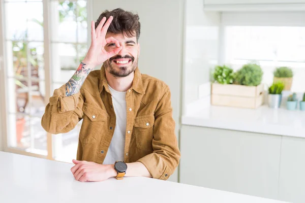 Young man wearing casual jacket sitting on white table doing ok gesture with hand smiling, eye looking through fingers with happy face.