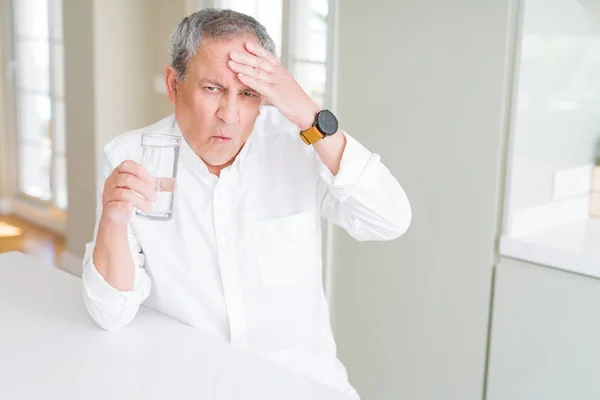 Handsome senior man drinking a fresh glass of water at home stressed with hand on head, shocked with shame and surprise face, angry and frustrated. Fear and upset for mistake.