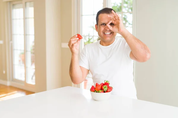 Middle age man eating strawberries at home with happy face smiling doing ok sign with hand on eye looking through fingers