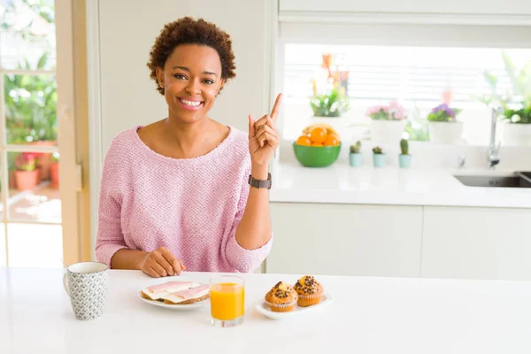 Young african american woman eating breaksfast in the morning at home with a big smile on face, pointing with hand and finger to the side looking at the camera.