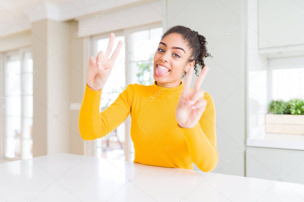 Beautiful african american woman with afro hair wearing a casual yellow sweater smiling with tongue out showing fingers of both hands doing victory sign. Number two.