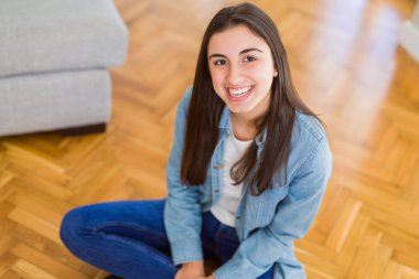 Güzel genç esmer kadın, arka planda güneş ışığı ile evde yerde oturan yüzünde büyük bir gülümseme ile kameraya bakarak neşeli gülümseyen