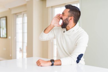 Yakışıklı İspanyol adam evde rahat beyaz kazak giyen bağırarak ve ağız da el ile yan yüksek sesle bağırıyor. İletişim kavramı.