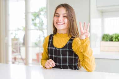 Genç, güzel sarışın kız evde sarı süveter giyiyor ve üç numaralı parmaklarıyla onu gösterirken kendinden emin ve mutlu gülümsüyor..