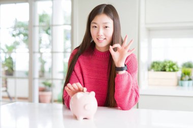 Güzel Asyalı kadın parmaklarıyla ok işareti yapıyor Piggy çanta içinde bir sikke koyarak, mükemmel sembolü