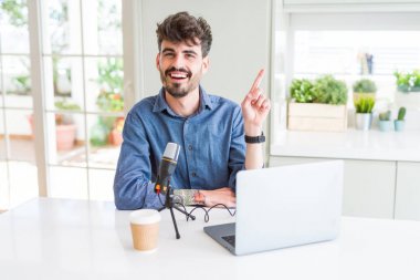 Yüz üzerinde büyük bir gülümseme ile mikrofon ve laptop kullanarak genç adam kayıt podcast, kamera bakarak yan el ve parmak ile işaret.