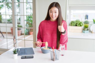 Güzel Asyalı kadın laptop kullanarak çalışan ve büyük bir gülümseme ile mutlu Asya noodle yeme ok işareti yapıyor, parmaklar ile başparmak, mükemmel işareti