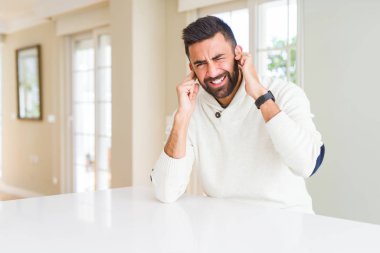 Yakışıklı İspanyol adam yüksek sesle müzik sesi için rahatsız ifade ile parmakları ile kulakları kapsayan evde rahat beyaz kazak giyiyor. Sağır kavramı.