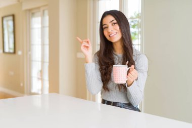 Bir fincan kahve içen genç bir kadın mutlu bir şekilde elini ve parmağını kenarı gösteriyor.