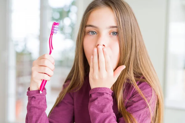 Beautiful young girl kid holding pink dental toothbrush cover mouth with hand shocked with shame for mistake, expression of fear, scared in silence, secret concept