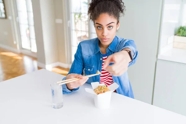 Young african american woman eating asian noodles from delivery box pointing with finger to the camera and to you, hand sign, positive and confident gesture from the front