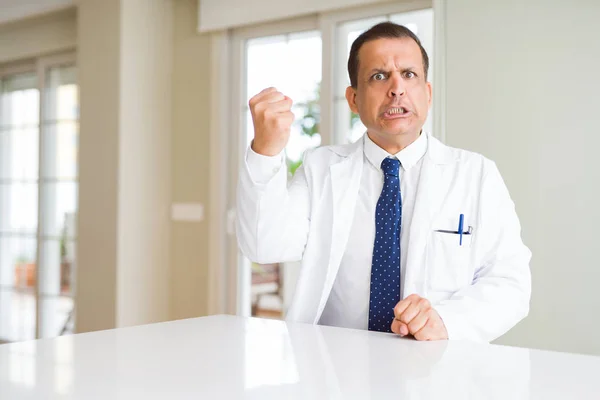 Middle age doctor man wearing medical coat at the clinic angry and mad raising fist frustrated and furious while shouting with anger. Rage and aggressive concept.