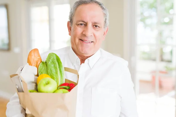 Handsome senior man holding paper bag full of fresh groceries and smiling at home