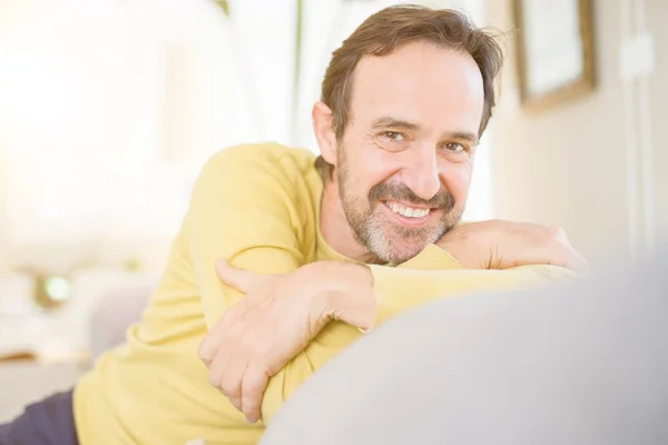 Handsome middle age man sitting on the sofa relaxed and smiling at the camera at home