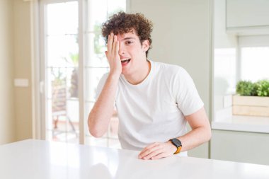 Evde beyaz tişört giyen yakışıklı genç bir adam bir gözünü kapatıyor, yüzünde kendine güvenen bir gülümseme ve sürpriz bir duygu var..