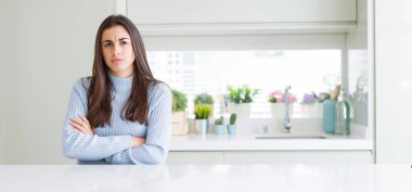 Geniş açı resim güzel genç kadının şüpheci ve sinir, onaylamayan ifade yüz çapraz kolları üzerinde evde beyaz masaya oturuyor. Negatif kişi.