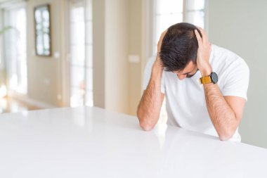 Yakışıklı İspanyol adam evde rahat beyaz t-shirt baş ağrısı umutsuz muzdarip ve stresli çünkü ağrı ve migren. Eller baş üstünde.