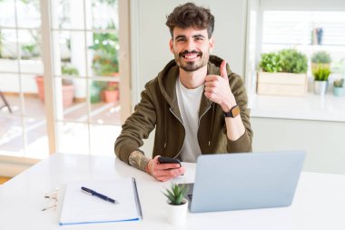 Akıllı telefon kullanarak genç öğrenci adam ve büyük gülümseme ile dizüstü mutlu kullanarak okuyan ok işareti yapıyor, parmaklar ile başparmak, mükemmel işareti