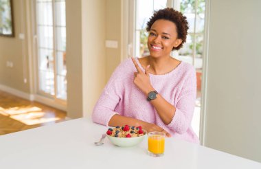 Genç Afro-Amerikalı kadın sabah sağlıklı bir kahvaltı yapıyor. Yüzünde mutlu ve doğal bir ifadeyle elleri ve parmaklarıyla gülümsüyor.
