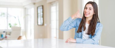 Evde beyaz masada oturan güzel genç kadının geniş açılı resmi telefonda konuşmak gibi el ve parmaklarla telefon hareketi yaparak gülümsüyor. İletişim kavramları.