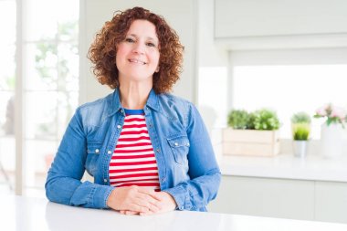 Kıvırcık saçlı orta yaş yaşlı kadın elleri birlikte evde kot ceket giyen ve rahat ve neşeli gülümseyen parmakları geçti. Başarı ve iyimser