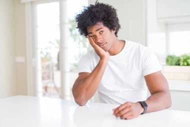 Beyaz bir tişört giyen genç Afro-Amerikan bir adam evinde oturmuş kollarını kavuşturup depresyondan sıkılmış ve yorgun göründüğünü düşünüyor..