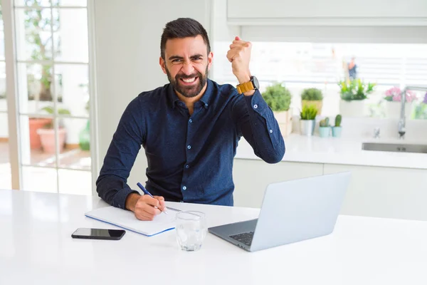 Handsome hispanic man working using computer and writing on a paper annoyed and frustrated shouting with anger, crazy and yelling with raised hand, anger concept