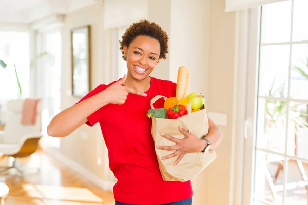 新鮮な食料品でいっぱいの紙袋を持つ若いアフリカ系アメリカ人女性は 手と指で指差し非常に幸せです — ストック写真
