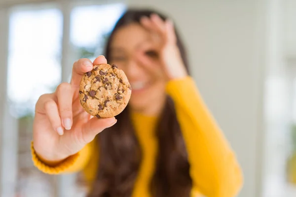 年轻女子吃巧克力片饼干在家里与快乐的脸微笑做确定的标志 手对眼睛看着通过手指 — 图库照片