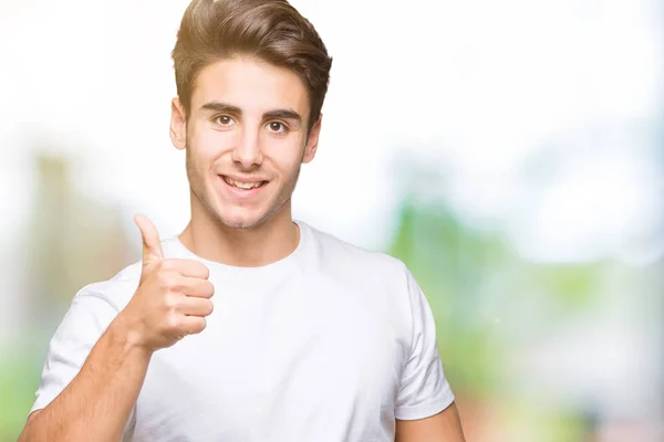 年轻英俊的男人穿着白色的 T恤在孤立的背景做快乐的大拇指用手的手势 查看显示成功的相机的审批表情 — 图库照片