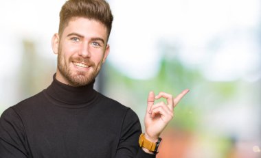 Kameraya bakarak yan el ve parmak ile işaret yüzünde, kocaman bir gülümseme ile kış kazak giyen genç sarışın yakışıklı.