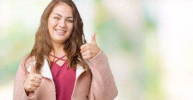 Artı boyutu genç kadın güzel izole arka plan başarı işareti el ile pozitif jest yapıyor üzerinde kış palto giyiyor başparmak gülümseyen ve mutlu. Neşeli bir ifadeyle, kazanan jest kameraya bakıyor.