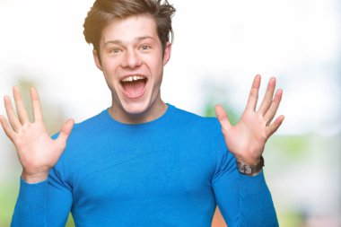 Üzerinde mavi kazak giyen genç yakışıklı adam deli kutluyor arka plan izole ve başarı için kaldırdı kollarını açarak hayrete ve heyecanlı çığlık gözleri açık. Kazanan kavramı