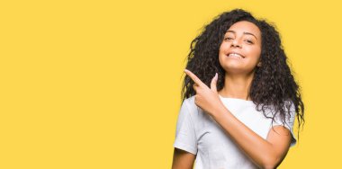 Kıvırcık saçlı beyaz t-shirt neşeli bir gülümseme ile el ve parmak kadar yan yüzünde mutlu ve doğal ifade ile işaret yüz ile rahat giyen genç güzel kız