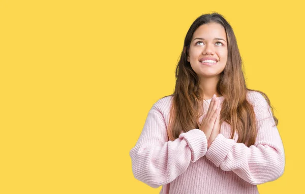 Young beautiful brunette woman wearing pink winter sweater over isolated background begging and praying with hands together with hope expression on face very emotional and worried. Asking for forgiveness. Religion concept.