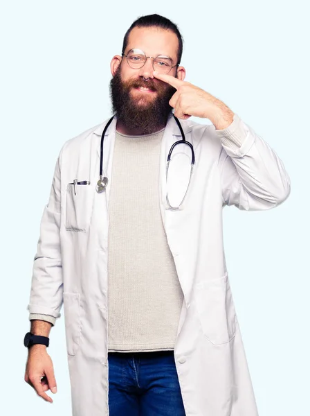 医療コートを着た若いブロンドの医者の男 顔と鼻に手指で指差し 陽気な笑顔 — ストック写真