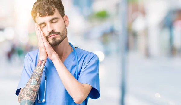 Young handsome nurse man wearing surgeon uniform over isolated background sleeping tired dreaming and posing with hands together while smiling with closed eyes.