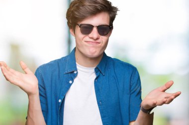 Genç yakışıklı adam izole arka plan clueless ve şaşkın bir ifadeyle kollar ve eller kaldırdı üzerinde güneş gözlüğü takıyor. Şüphe kavramı.