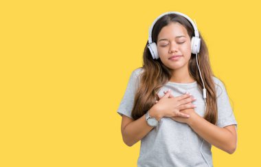 Genç güzel kadın kulaklıklar kapalı gözler ve yüz minnettar jest ile göğsüne eller ile gülümseyen izole arka plan üzerinde müzik dinlerken giyiyor. Sağlık konsepti.