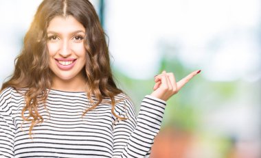 Yüzünde büyük bir gülümseme ile çizgili kazak giyen genç güzel kadın, kameraya bakarak yan el ve parmak ile işaret.