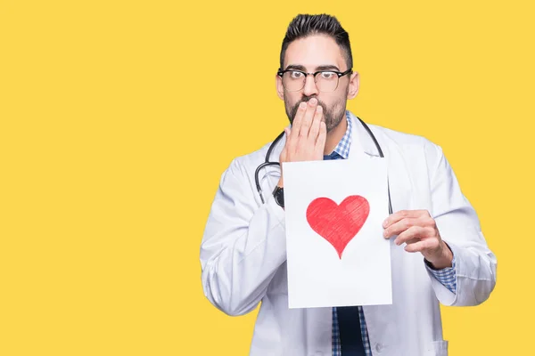 Handsome young doctor man holding paper with red heart over isolated background cover mouth with hand shocked with shame for mistake, expression of fear, scared in silence, secret concept