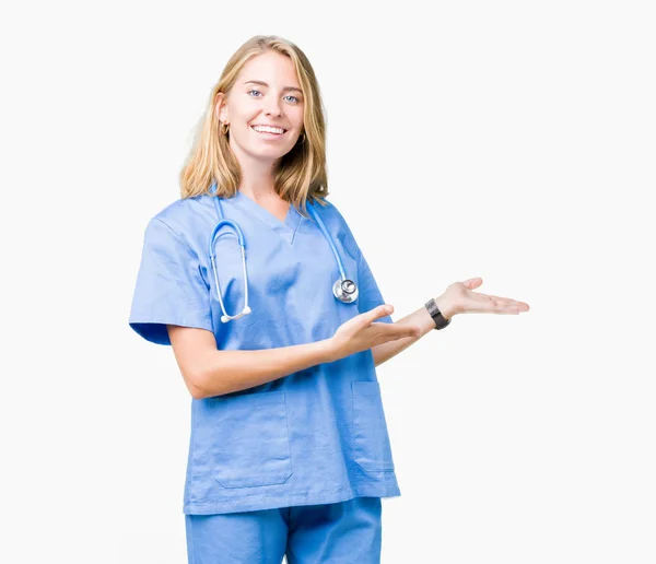 美丽的年轻医生妇女穿着医疗制服在孤立的背景邀请进入微笑自然与开放的手 — 图库照片