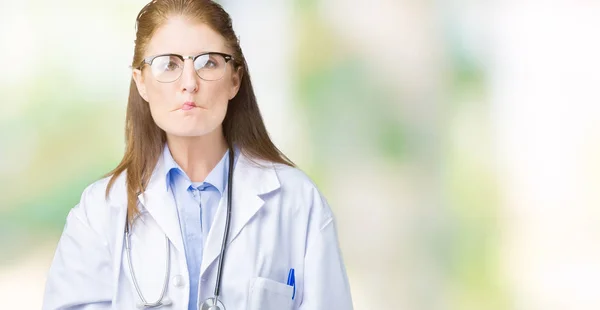 Middle age mature doctor woman wearing medical coat over isolated background making fish face with lips, crazy and comical gesture. Funny expression.